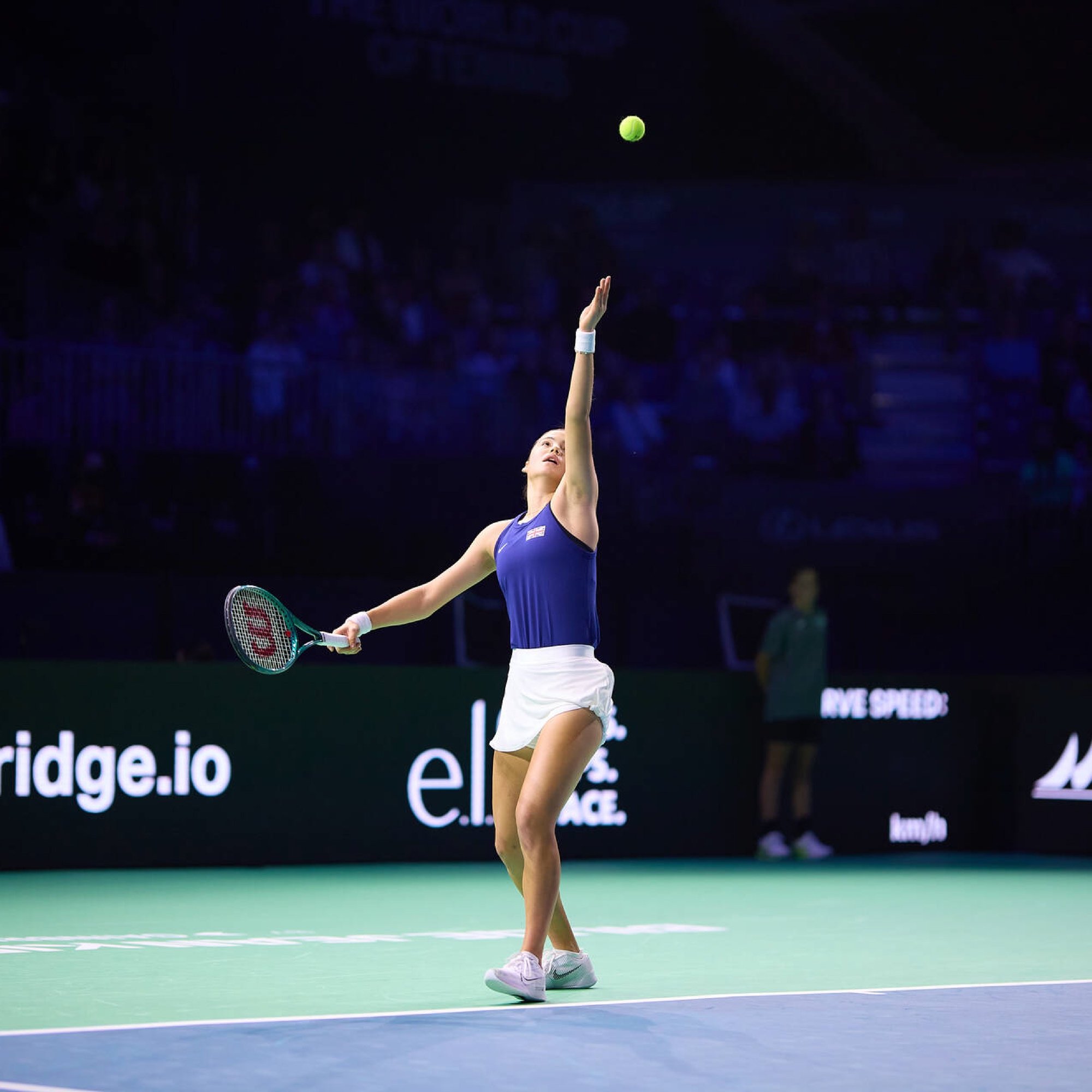 Tennis player in a blue top and white skirt serves on a court, with a ball in the air.