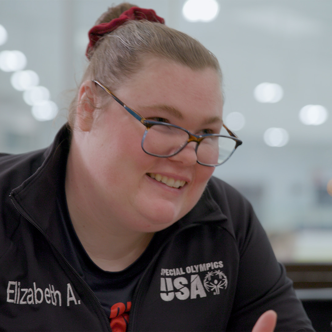 A woman wearing glasses and a "Special Olympics USA" jacket with the name "Elizabeth A." smiles indoors.
