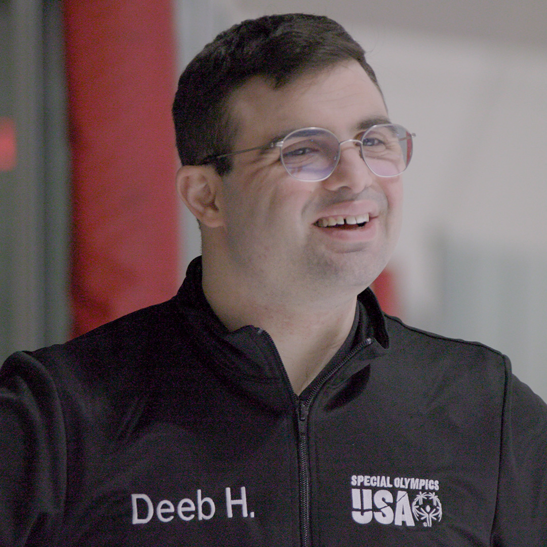 A man wearing glasses and a black Special Olympics USA jacket with the name "Deeb H." stands indoors, smiling.