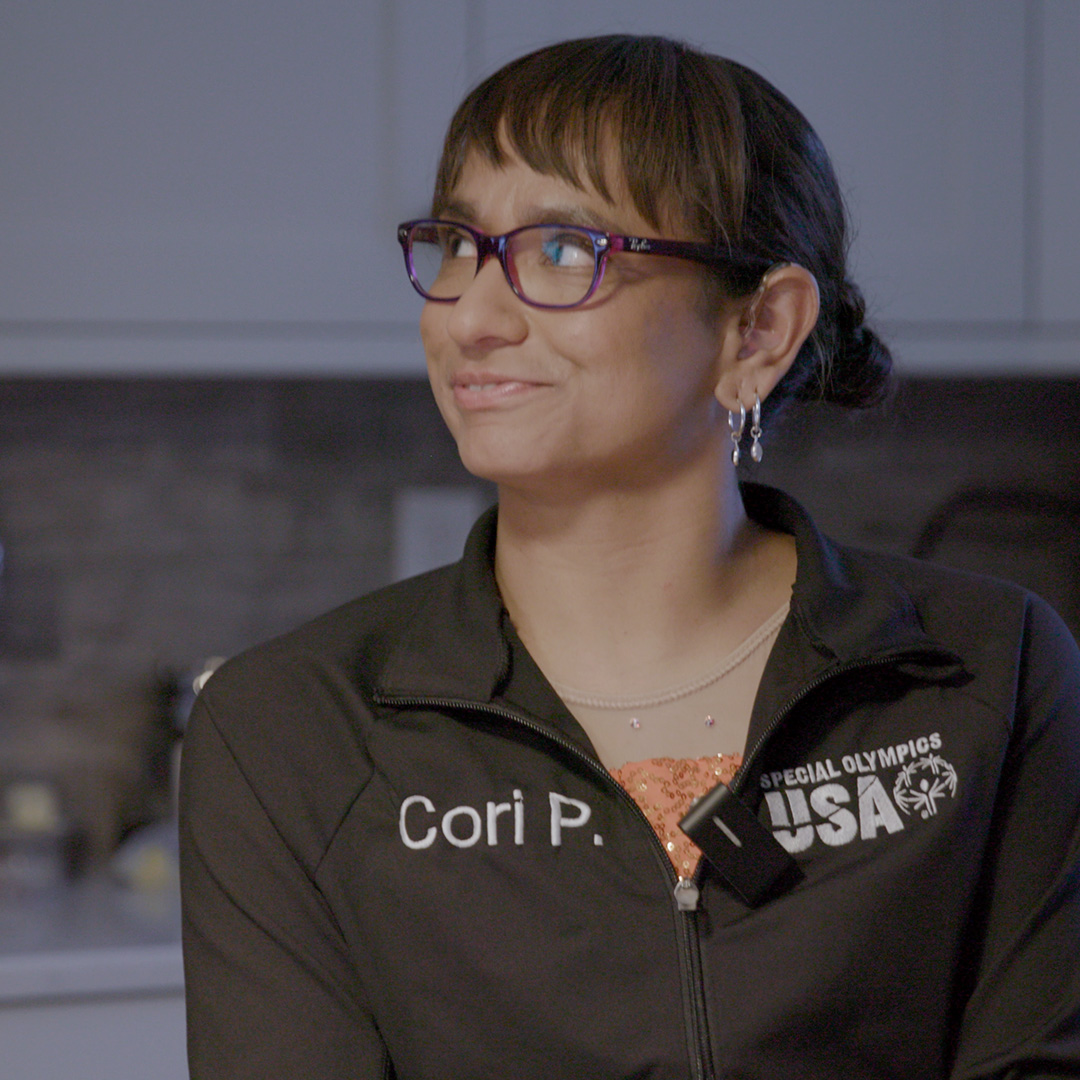 A woman wearing glasses and a "Special Olympics USA" jacket with the name "Cori P." looks to the side in a kitchen setting.