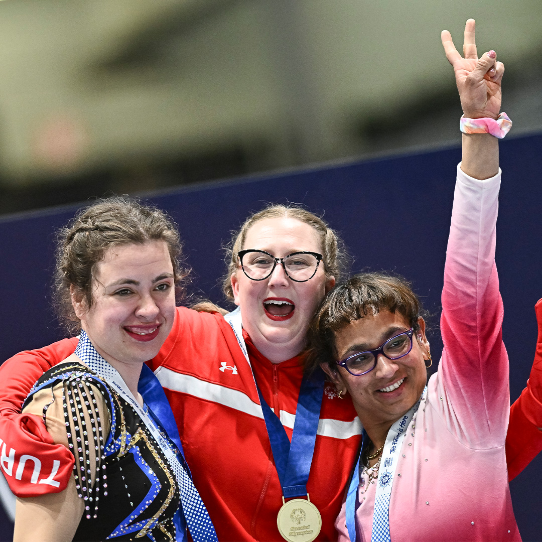 Three female athletes wearing medals smile and pose together; the athlete on the right raises her hand in a peace sign.