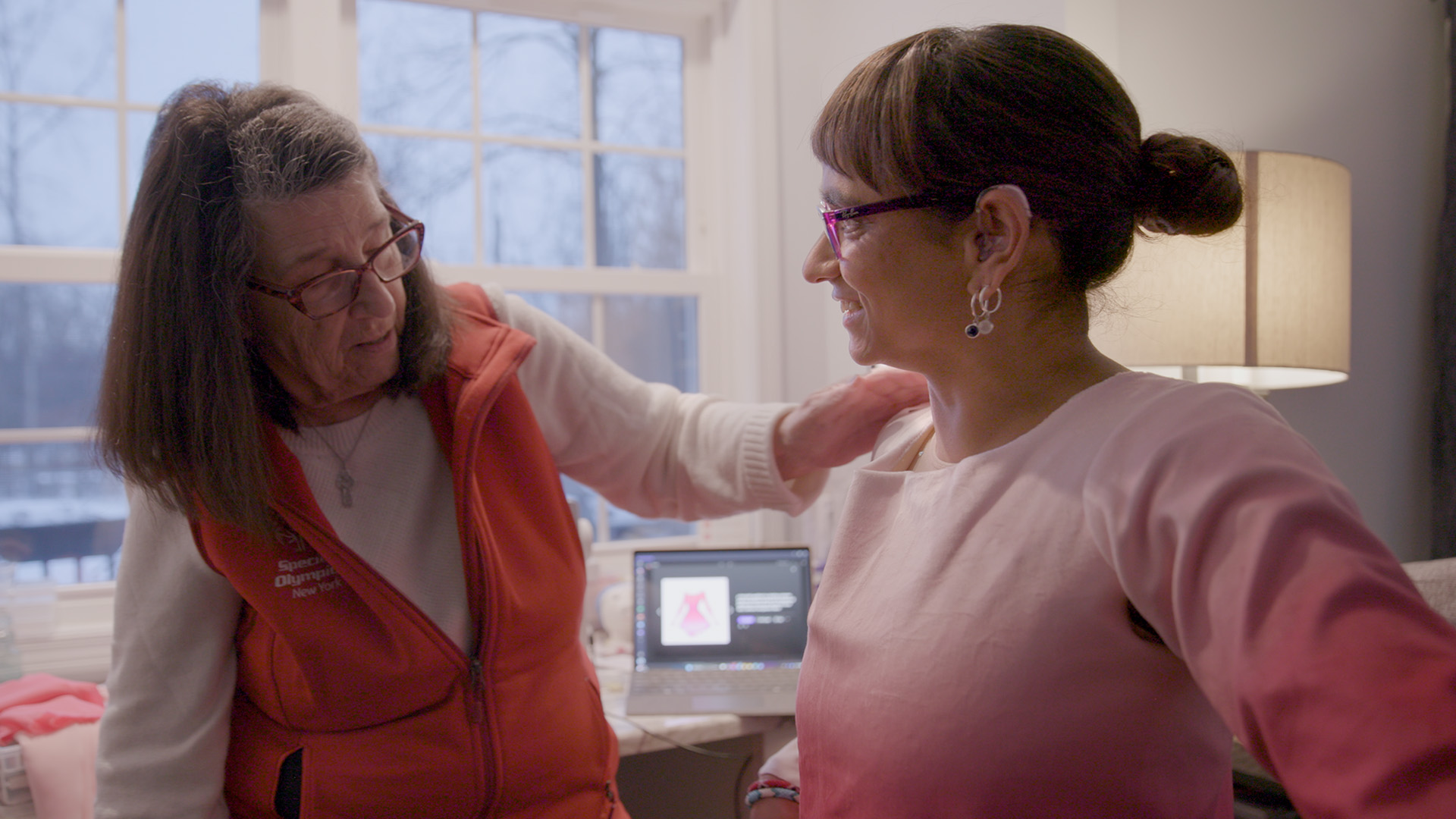 An older woman places a comforting hand on a younger woman's shoulder as they talk in a bright room with a laptop on the desk behind them.