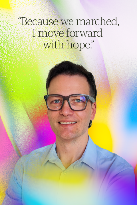 A person wearing glasses and a light blue shirt smiles in front of a colorful background with the quote, "Because we marched, I move forward with hope.