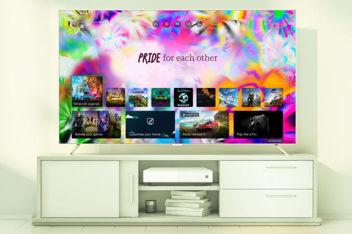 A TV displays a colorful Pride-themed screen with the text "PRIDE for each other," showing various video game icons. The TV sits on a white entertainment unit with a console and speakers.